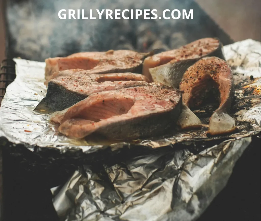 Freshly grilled salmon steaks on aluminum foil over an open flame, seasoned with spices and herbs.