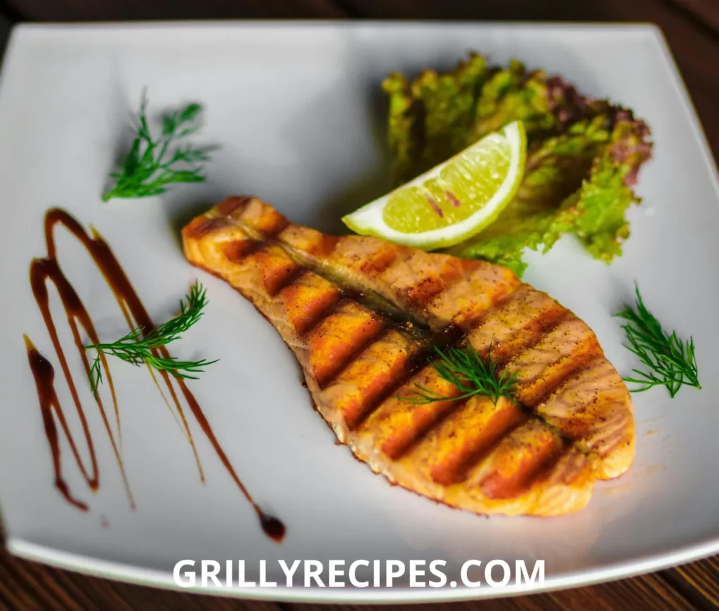 Grilled salmon steak with grill marks, garnished with fresh dill, lettuce, and a lemon wedge on a white plate.