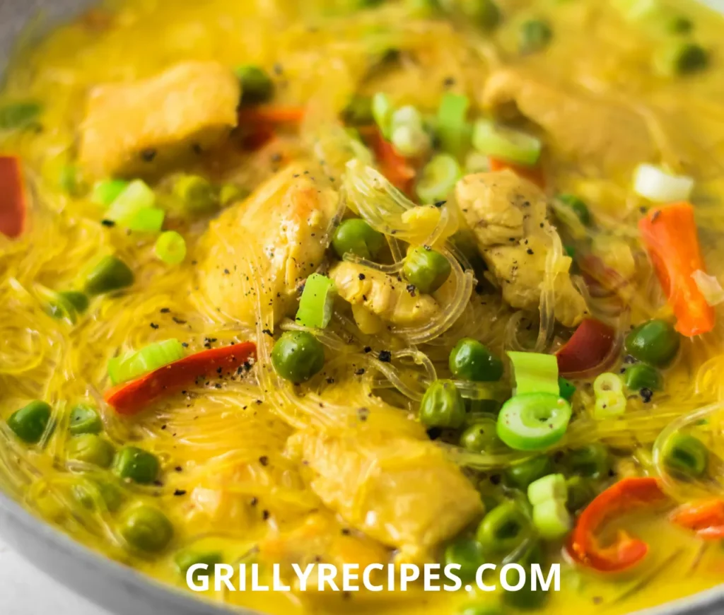A bowl of golden noodle soup with glass noodles, chicken, green peas, and red peppers, garnished with black pepper and green onions.